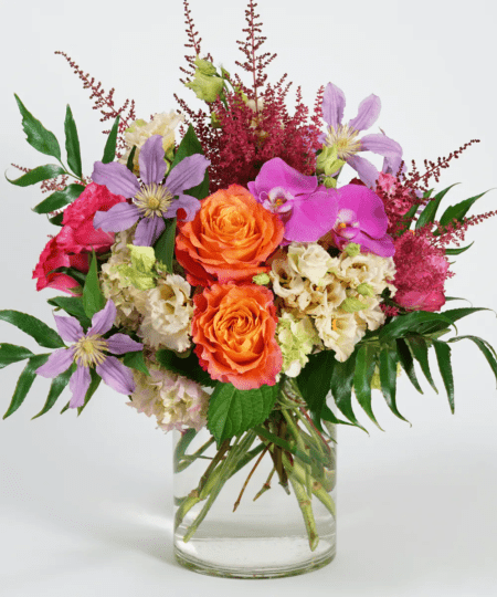 This unique design features a picturesque collection of jewel toned blooms including orange roses, fuchsia orchids, and lavender clematis accented with burgundy astilbe and holly fern and is designed in a glass cylinder vase.