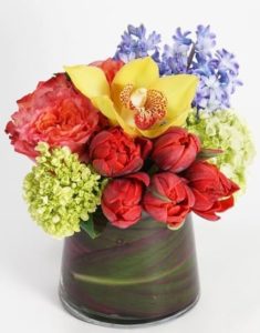 A rainbow of color, this petite masterpiece includes tulips, roses, and hyacinth topped with a bright yellow cymbidium - all gathered in a unique glass vase lined with a burgundy ti leaf