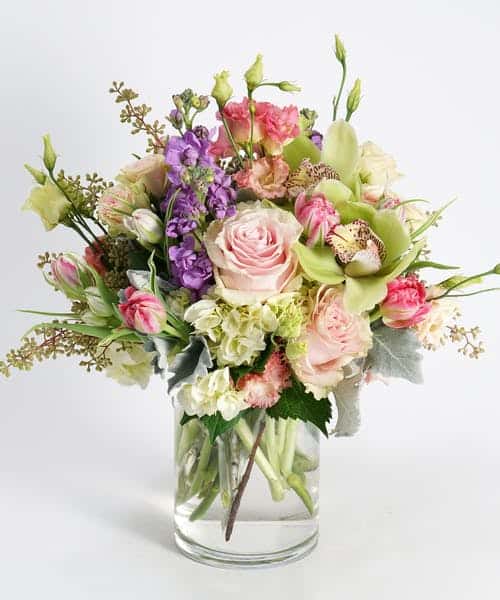 This gardeny style design features a beautiful bevy of pretty, pastel toned blooms including soft peach roses, pink tulips, lavender stock, white hydrangea and green cymbidium orchids arranged in our signature glass cylinder vase.