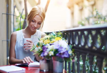 Attractive young girl reading book while drinking coffee at sunny day sitting on the balcony